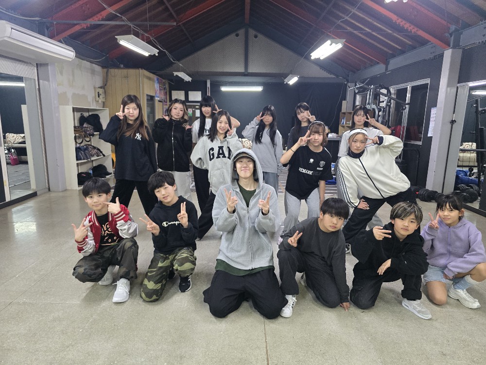 ダンススタジオでピースサインをしてポーズをとる、カジュアルな服装の若者と子供たちの集合写真。